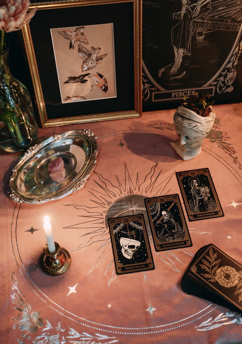 May include: A tarot card reading spread with four cards laid out on a pink table cloth with a silver sun and moon design. The cards are "Knight of Cups", "Queen of Swords", "Page of Wands", and "Page of Death".