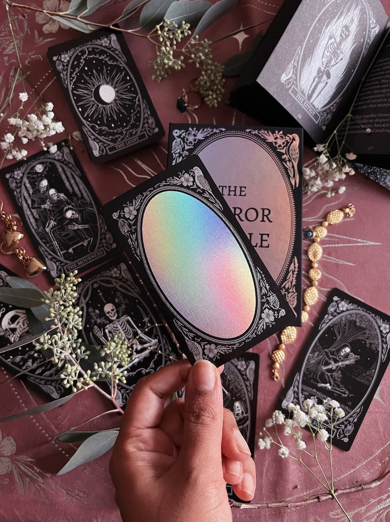 Peut inclure: Une carte de tarot noire avec un ovale holographique iridescent au centre. La carte est entour&eacute;e d'une bordure d&eacute;corative avec des fleurs et des oiseaux. Le texte "The Mirror Oracle" est imprim&eacute; sur la carte.