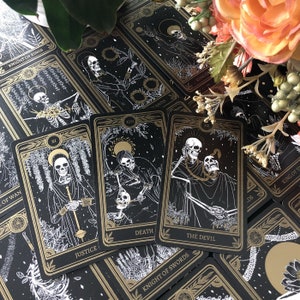 May include: A deck of tarot cards with black backgrounds and gold and white illustrations. Three cards are in focus, labeled "Justice", "Death", and "The Devil". Other cards are visible, including "Knight of Swords" and "Knight of Rings". Floral accents frame the cards.