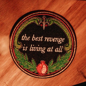 Peut inclure: Un écusson brodé noir et vert avec une grenade rouge et le texte "the best revenge is living at all".