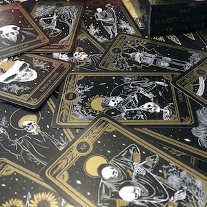 May include: A collection of tarot cards with black backgrounds and gold and white illustrations. The cards feature images of skeletons, celestial bodies, and botanical elements. The text on the cards includes "The Star," "King of Cups," and "Death."