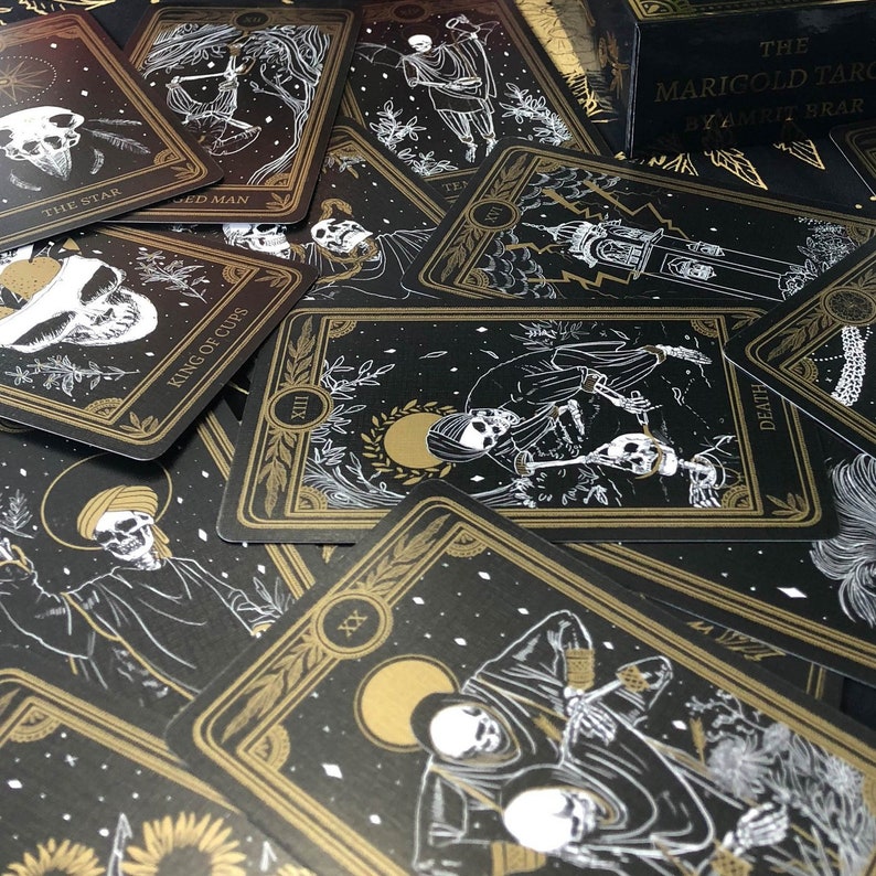 May include: A collection of tarot cards with black backgrounds and gold and white illustrations. The cards feature images of skeletons, celestial bodies, and botanical elements. The text on the cards includes "The Star," "King of Cups," and "Death."