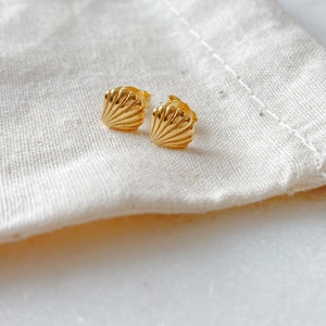 May include: A pair of gold-toned stud earrings shaped like small seashells. Each earring features a fan-like design with raised ridges radiating from the base to the scalloped edge, finished with a polished, reflective surface and standard earring backs.