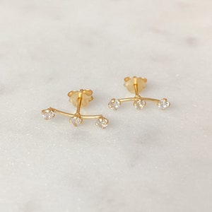 May include: A pair of gold earrings with three small clear stones on each earring. The earrings are curved and have a post back.
