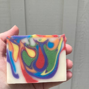 以下が含まれることがあります： 赤、黄、緑、青、紫の色合いでカラフルな渦巻き模様が施された手作り石鹸。石鹸は白く、滑らかで磨かれた表面をしています。