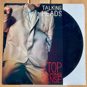 May include: A vinyl record cover for Talking Heads Stop Making Sense featuring a person wearing an oversized light beige suit jacket. The text Talking Heads appears in white at the top right, and Stop Making Sense is printed in red at the bottom.