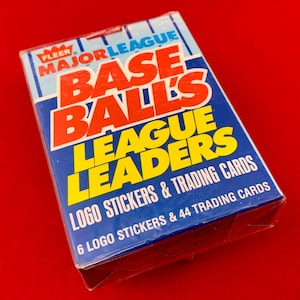 May include: A vintage unopened pack of Fleer Major League Baseball League Leaders trading cards. The box is blue with red and yellow text, including "BASEBALLS LEAGUE LEADERS" and "LOGO STICKERS & TRADING CARDS". The background is a solid red.