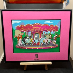 May include: Framed print featuring a carousel scene with Looney Tunes characters at Six Flags. The image has a pink mat and a black frame. The carousel is a vibrant pink and gold, with Bugs Bunny and other characters riding.