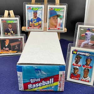 May include: A collection of baseball cards, including rookie cards and top prospects, displayed in protective sleeves. A box of 1992 Topps Major League Baseball cards is in the foreground. The cards feature various players and team logos.
