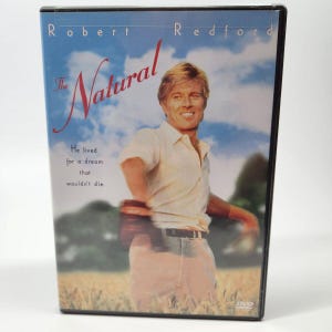 May include: DVD case for the movie "The Natural" featuring Robert Redford. The cover art shows Redford in a white shirt and tan pants against a backdrop of a wheat field and blue sky. Text includes the movie title and a quote.