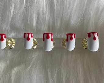 May include: Five white artificial nails with a red dripping blood design. The nails are displayed on a clear plastic strip with gold-colored hardware, set against a fluffy white background.