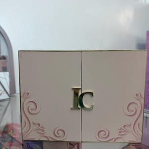 Könnte beinhalten: Eine hellrosa Schachtel mit goldenen Rändern und einem goldenen 'IC'-Emblem. Die Schachtel hat ein dekoratives Wirbeldesign. Die Schachtel ist geöffnet und gibt den Blick ins Innere frei.