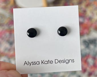 Puede incluir: Un par de pendientes de botón pequeños, redondos y de color negro brillante montados en una tarjeta de presentación rectangular blanca. La tarjeta presenta el texto impreso en negro Alyssa Kate Designs en la parte inferior. Tienen una superficie lisa.