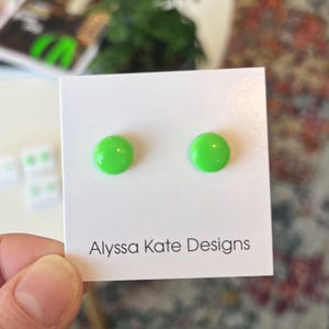 May include: Two bright green, circular stud earrings displayed on a white card. The card features the text "Alyssa Kate Designs" in black. The earrings are a vivid, solid colour.