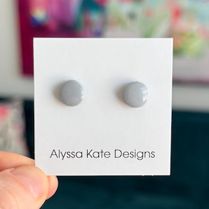 May include: Two light gray, round stud earrings displayed on a white card. The card has the text "Alyssa Kate Designs" in a black, simple font. The earrings are glossy and appear to be made of a smooth material.