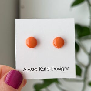 May include: A pair of bright orange stud earrings on a white card. The earrings are round and glossy. The card has the text "Alyssa Kate Designs" in a black, simple font.