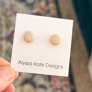 May include: A pair of small, round, beige stud earrings on a white card. The card has the text "Alyssa Kate Designs" printed in black. The earrings are set against a patterned background.