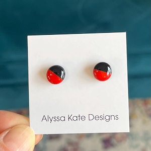 May include: A pair of round stud earrings. Each earring is half black and half red. The earrings are displayed on a white card with the text "Alyssa Kate Designs" in black.