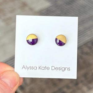 May include: A pair of round stud earrings, each with a gold upper half and a deep purple lower half. The earrings are displayed on a white card with the text "Alyssa Kate Designs".