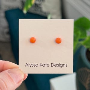 May include: A pair of small, round, bright orange stud earrings attached to a square white display card. The card features the black printed text Alyssa Kate Designs at the bottom. The earrings have a smooth, glossy finish and a simple circular shape.