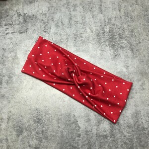 May include: A red headband with white polka dots. The fabric is twisted at the centre, creating a stylish accessory. The headband is made of a soft material and is suitable for various hairstyles.