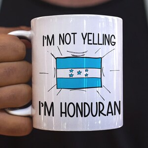 May include: White ceramic mug with the text "I'M NOT YELLING I'M HONDURAN" and a graphic of the Honduran flag. The mug has a white handle and is held by a person.