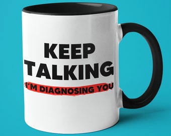 May include: White ceramic coffee mug with a black rim and handle. The mug has black text that reads "KEEP TALKING I'M DIAGNOSING YOU" with a red line through the "I'M DIAGNOSING YOU" portion.