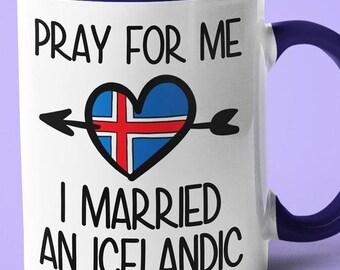 May include: White coffee mug with a blue rim and handle. The mug has a black and white graphic that says "Pray for me I married an Icelandic" with a heart shaped Icelandic flag.