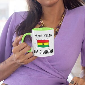 May include: White ceramic mug with a green rim and handle. The mug has a black and white graphic of the flag of Ghana with the text "I'm not yelling I'm Ghanaian".