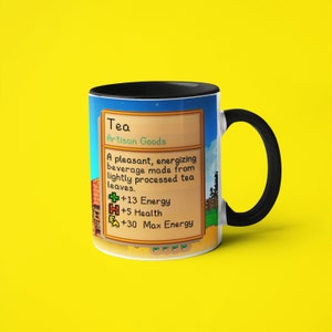 Op de afbeelding: Een witte keramische mok met een zwart handvat heeft een gepixelde afbeelding van een videogame-item genaamd "Thee". De tekst luidt "Ambachtelijke goederen" en beschrijft het item als "Een aangenaam, energiek drankje gemaakt van licht verwerkte theebladeren." De afbeelding toont ook de statistieken van het item: +13 Energie, +5 Gezondheid en +30 Maximale Energie.
