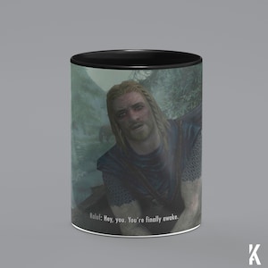 Op de afbeelding: Een zwart-witte mok met een sc&egrave;ne uit de videogame "The Elder Scrolls V: Skyrim". De mok toont een personage uit het spel, Ralof, met de tekst "Ralof: Hey, you. You're finally awake." gedrukt op de bodem.