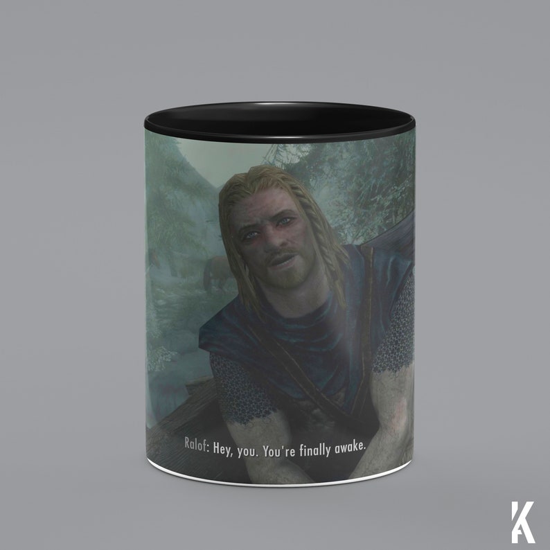 Op de afbeelding: Een zwart-witte mok met een sc&egrave;ne uit de videogame "The Elder Scrolls V: Skyrim". De mok toont een personage uit het spel, Ralof, met de tekst "Ralof: Hey, you. You're finally awake." gedrukt op de bodem.