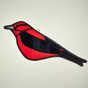 May include: A stained glass bird in a vibrant red and black color scheme. The bird features a detailed design with a black outline and a small metal loop for hanging. The bird's body is primarily red, with black wings and tail.