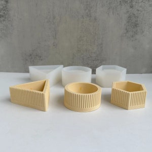 May include: Five different shaped silicone moulds for making candles or soap. The moulds are in various geometric shapes, including a triangle, circle, hexagon, and two different sized rectangles. The moulds are white and have a smooth, glossy finish.