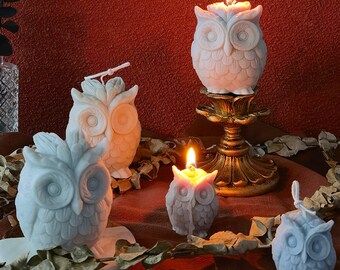 May include: Four white owl-shaped candles with a flame burning on top of each. The candles are on a wooden surface with green leaves and a gold metal stand.