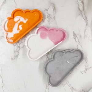 Puede incluir: Tres bandejas decorativas en forma de nube de diferentes colores. La primera bandeja es naranja con remolinos blancos, la segunda es blanca con remolinos rosas y la tercera es gris.