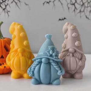 Könnte beinhalten: Eine Sammlung farbenfroher Zwergfiguren, perfekt für Halloween. Die Zwerge sind in Gelb-, Blau- und Beigetönen gehalten, mit kürbisförmigen Körpern und Hüten. Einer ist ein geschnitzter Kürbis. Der Hintergrund ist hellgrau.