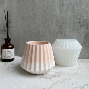Può includere: Due vasi in cemento con un designo a coste. Il vaso a sinistra è di colore rosa chiaro con una base bianca. Il vaso a destra è bianco.