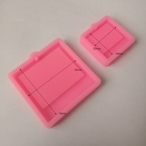 May include: Two pink silicone molds for crafting. The larger mold is square, measuring 50mm x 50mm, with a rectangular interior. The smaller mold is also square, 30mm x 30mm, with a rectangular interior. Both have a loop for hanging.