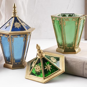 May include: Decorative glass containers with gold trim. One is blue with a lid, another is green, and the lid is off. The lids and containers have sun and star designs. The containers are on a light surface.
