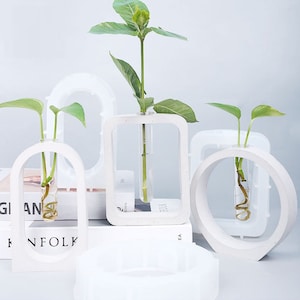 May include: Four white concrete vases with different geometric shapes, each with a glass test tube for holding small plants. The vases are arranged on a white surface with a book in the background.