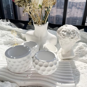 May include: White ceramic planters with a textured surface, a white ceramic vase with dried flowers, and a white ceramic bust of a person. The planters and vase are sitting on a white surface.