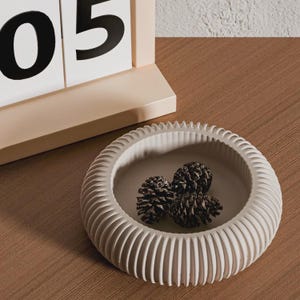 May include: A cream-coloured, ribbed ceramic bowl contains three pine cones. The bowl is on a wooden surface, with a beige calendar in the background showing the number "05".
