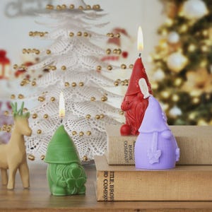 Könnte beinhalten: Drei zwergenförmige Kerzen in Grün, Rot und Lila, mit brennenden Dochten. Die Kerzen stehen auf Büchern, mit einem weißen Weihnachtsbaum aus Spitze und einer Rentierfigur im Hintergrund. Eine festliche Weihnachtsdekoration.