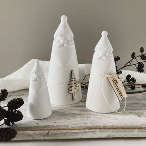 Könnte beinhalten: Drei weiße Keramik-Weihnachtsmannfiguren in verschiedenen Größen. Der größte hat einen Holzbaum, und der mittlere hat ein Etikett mit der Aufschrift "Weihnachten". Die Figuren stehen auf einem Holztablett mit Dekorationselementen.