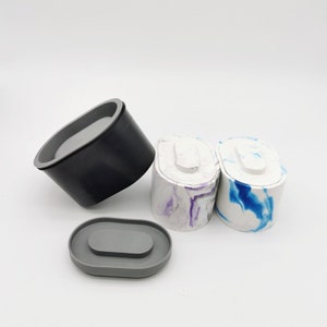 Puede incluir: Tres contenedores de hormigón de forma ovalada con tapas. Dos contenedores tienen una base blanca con vetas azules y moradas. El tercer contenedor es negro. Las tapas son grises.