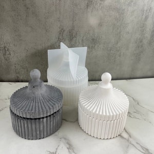 Può includere: Tre barattoli per candele decorativi in ​​calcestruzzo con coperchi. I barattoli sono di colore grigio, bianco e bianco traslucido. I barattoli hanno un design scanalato e una manopola rotonda sul coperchio.