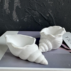 Op de afbeelding: Twee witte keramische schelpvormige plantenpotten. De potten zijn gevormd als schelpen en hebben een glad, getextureerd oppervlak.