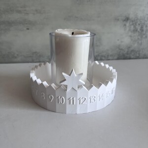 May include: White candle holder with a star accent and a clear glass cylinder. The holder has a scalloped edge with numbers 7 through 15. A white candle sits inside the glass.