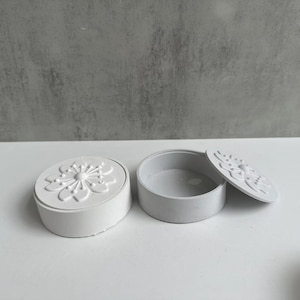 May include: A white, round, decorative trinket box with a floral design on the lid. The lid is slightly ajar, revealing the interior of the box. The box is made of a smooth, matte material.